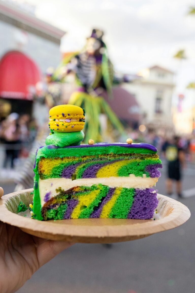 Vibrant slice of Mardi Gras cake with macaron topping, held at festive outdoor event.