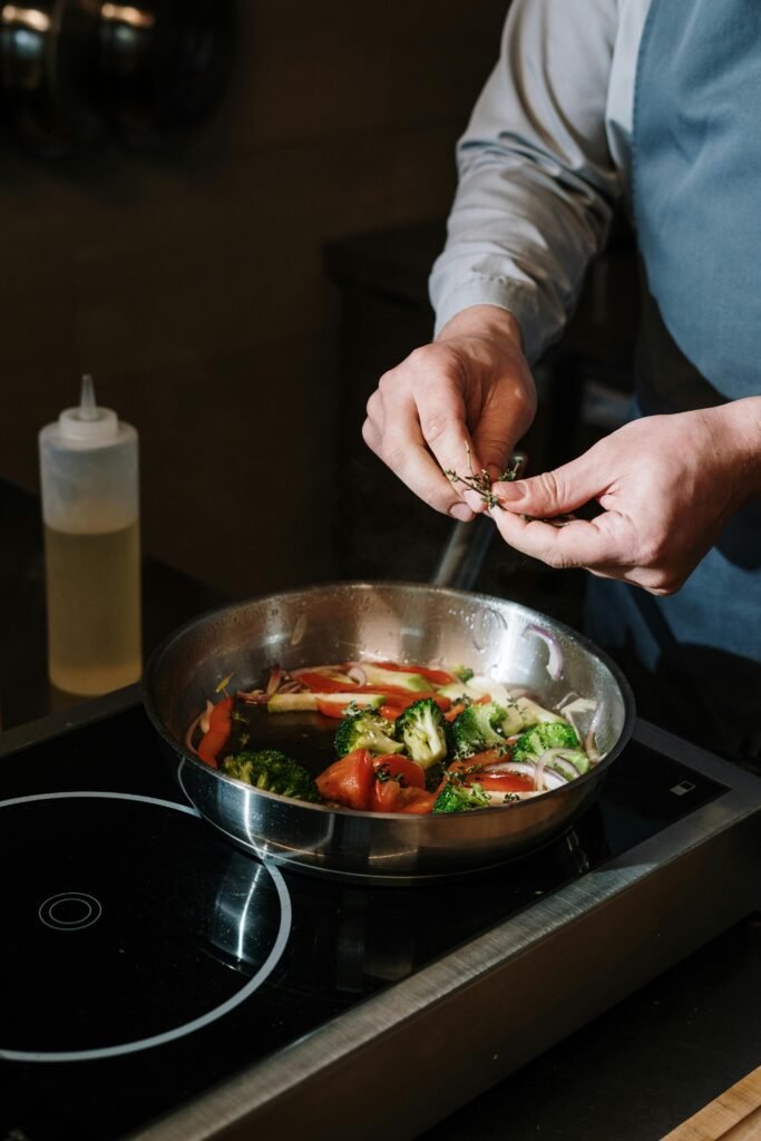 A chef seasons vegetables in a frying pan on a modern stovetop indoors.